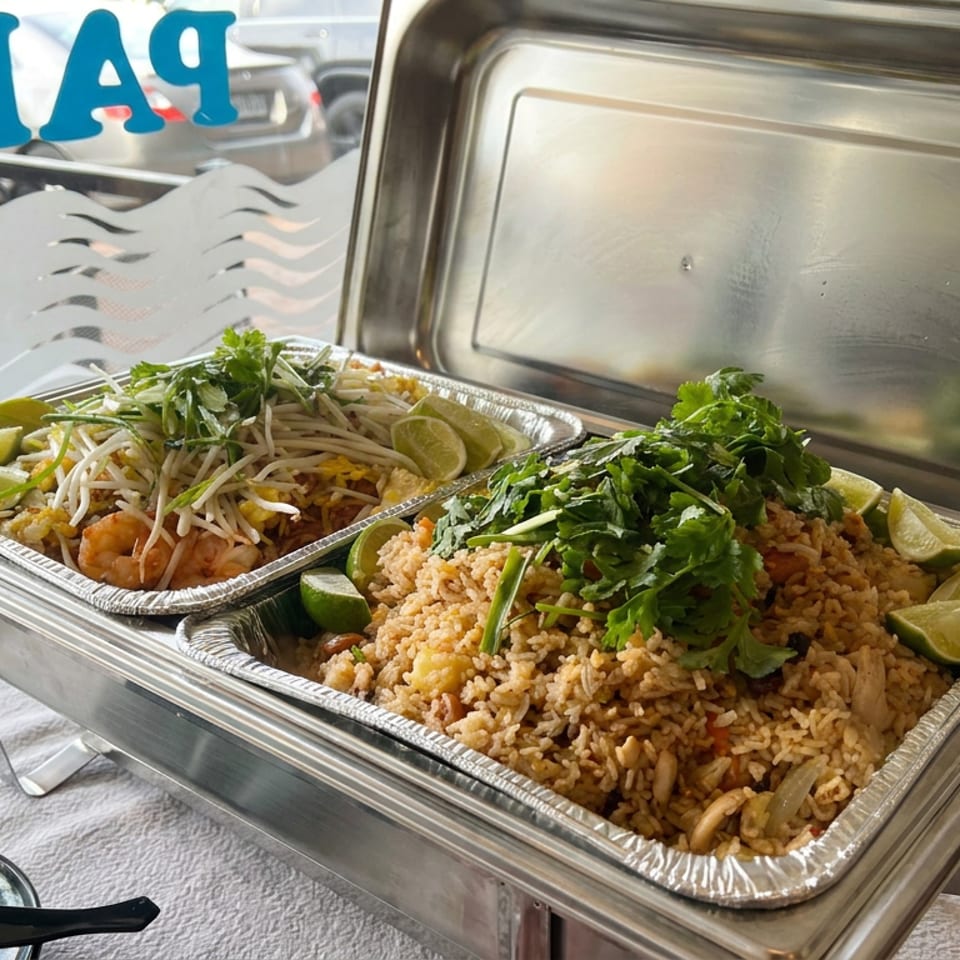 Two aluminum trays hold fried rice and pad Thai, garnished with cilantro, bean sprouts, and lime wedges.