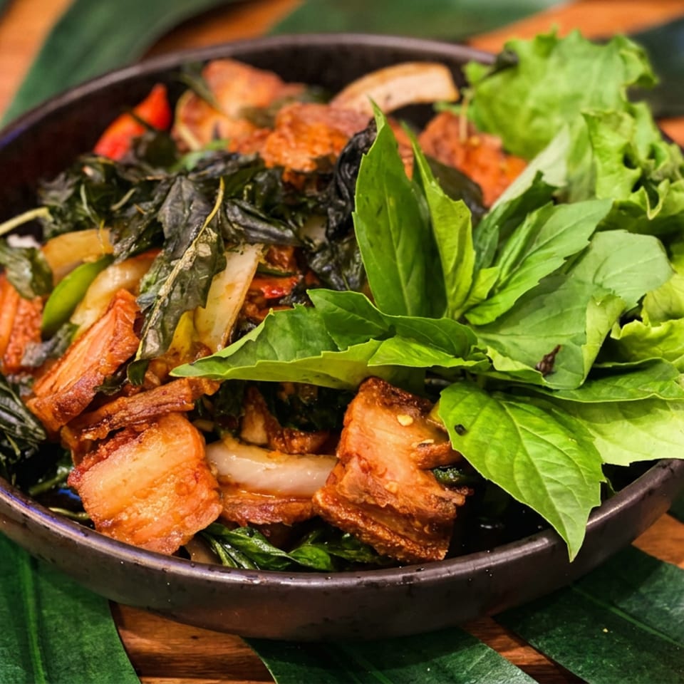 Bowl of crispy pork, sautéed vegetables, herbs, and fresh lettuce on a dark plate atop tropical leaves.