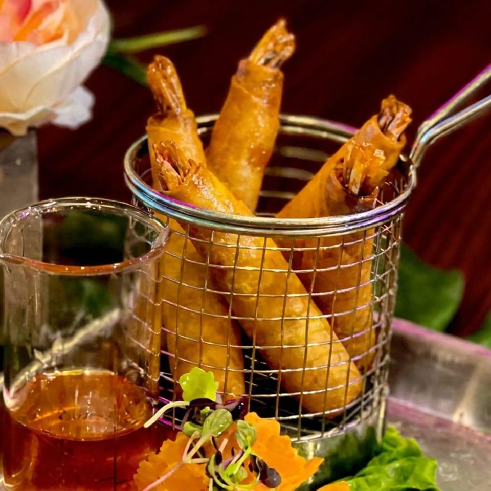 Golden fried shrimp served in a wire basket with dipping sauce, microgreens, and floral garnish on a metal tray.